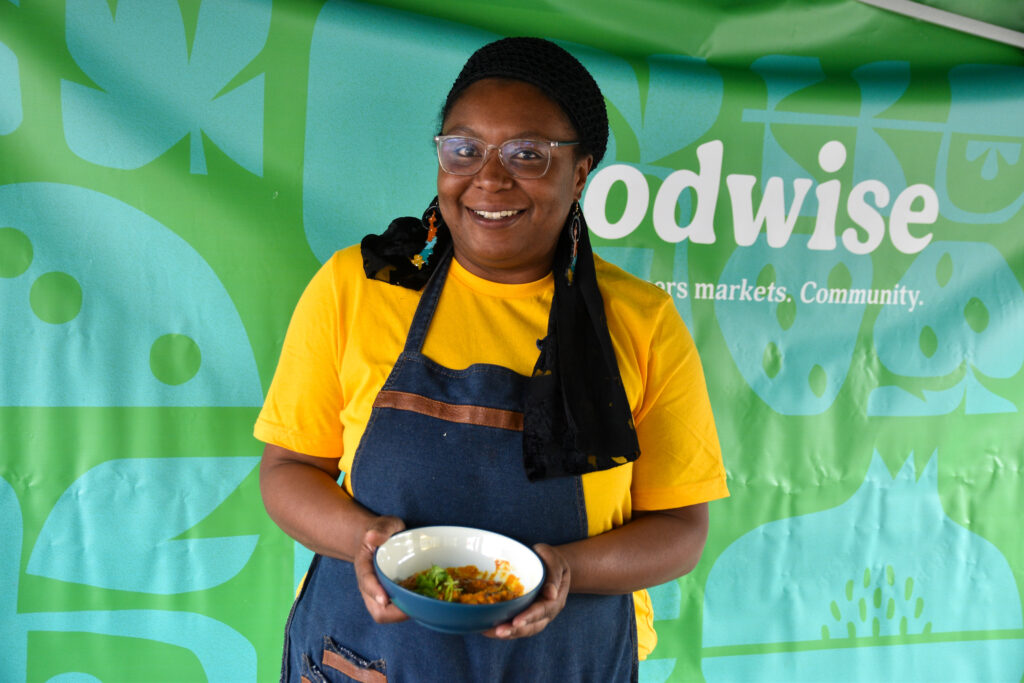 Angélica Mena poses with a bowl of Encocado de Pescado del Pacifico (Coconut Fish from the Colombian Pacific) in front of a green and blue backdrop at the Foodwise Classroom at the Ferry Plaza Farmers Market in San Francisco.