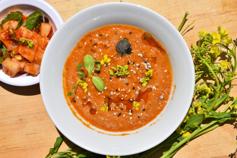 A bowl of bright orange-red kimchi gazpacho, served alongside kimchi and broccoli flowers